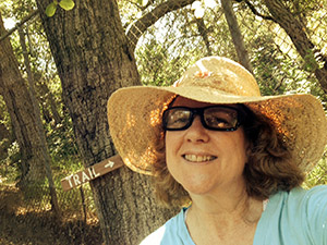 Laurie in a straw sunhat by a tree marked with a trail sign.