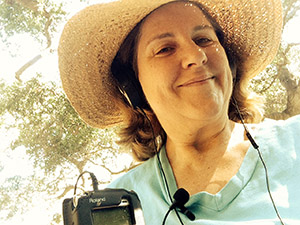 Laurie with two mics wearing headphones along with her floppy hat under the trees.