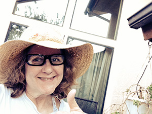 Laurie shows thumbs up in a straw hat and glasses in front of the windows of her house.