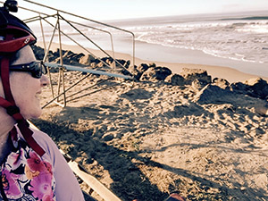 Laurie in bike gear looking out at waves on a beach