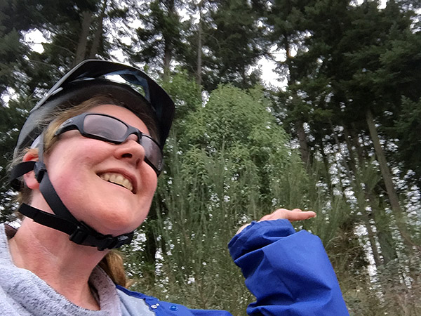 Laurie in bike gear and raincoat standing in front of evergreen trees