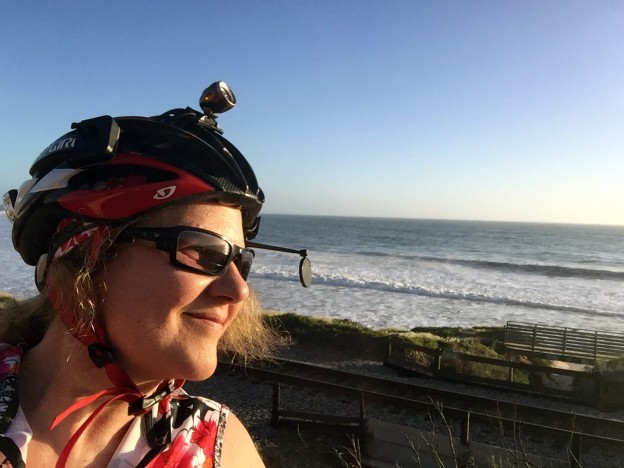 Laurie in bike gear looks out to sea with rolling waves on a sunny day.