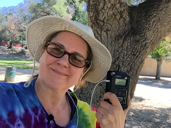Laurie in a rainbow tie-dye jacket and hiking hat standing under a tree with her recorder.