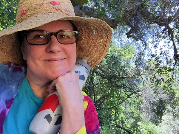 Laurie in her colorful jacket sitting under a tree with her straw hat and water bottle