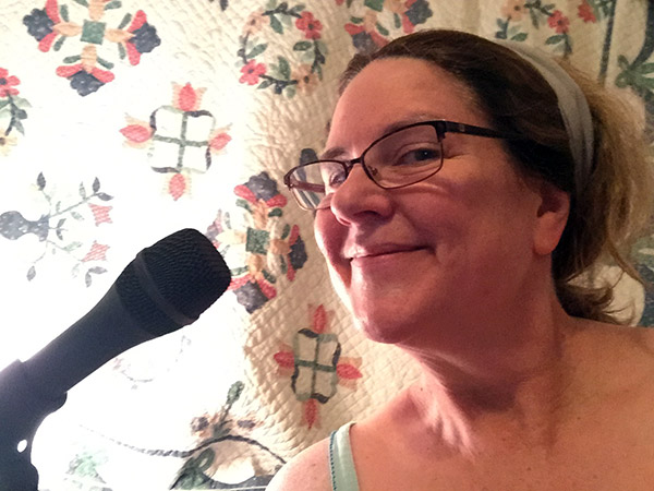 Laurie wearing her P.J.s by the mic in front of a quilt hung over the window