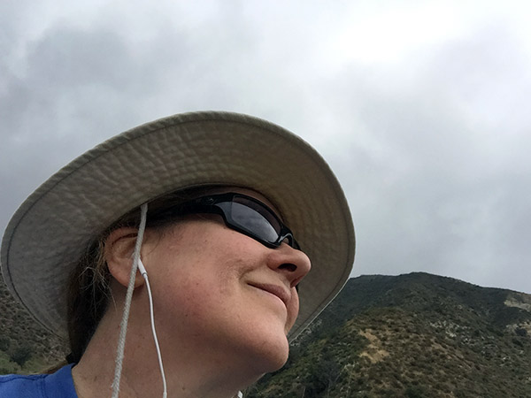 Shot looking at the mountain and sky from below Laurie's chin. She's wearing sun glasses and her hiking hat.