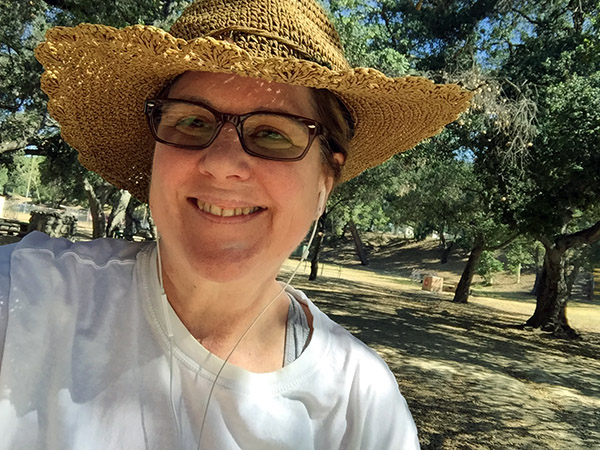 Laurie in her fancy woven sunhat smiles under trees in the park.