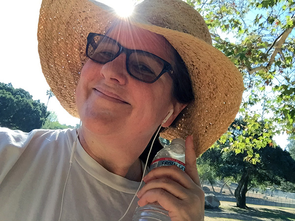 Laurie in her straw hat in a ray of sunshine holding a bottle of water at the park.