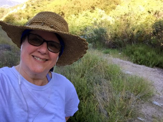 Laurie smiling in her straw hat with the sun shining down on the hiking trail.