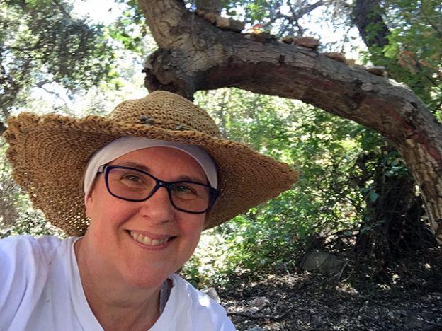 Laurie wearing a white shirt and sunhat smiles under a tree