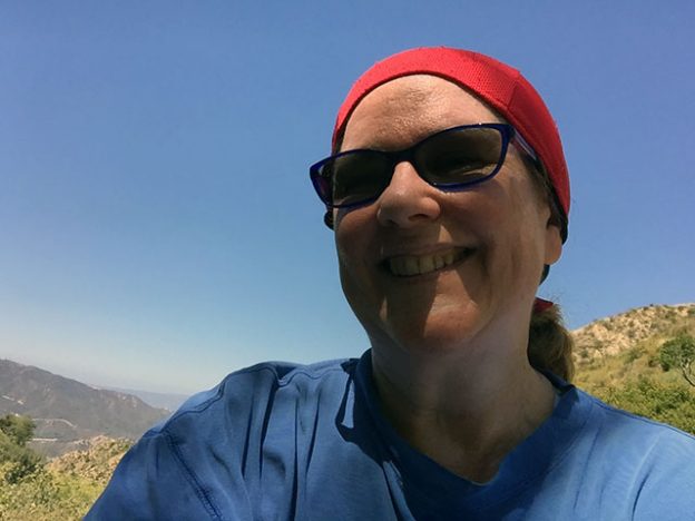 Laurie in red head scarf and blue long sleeved shirt at top of trail with bright cloudless sky behind - face in shadow