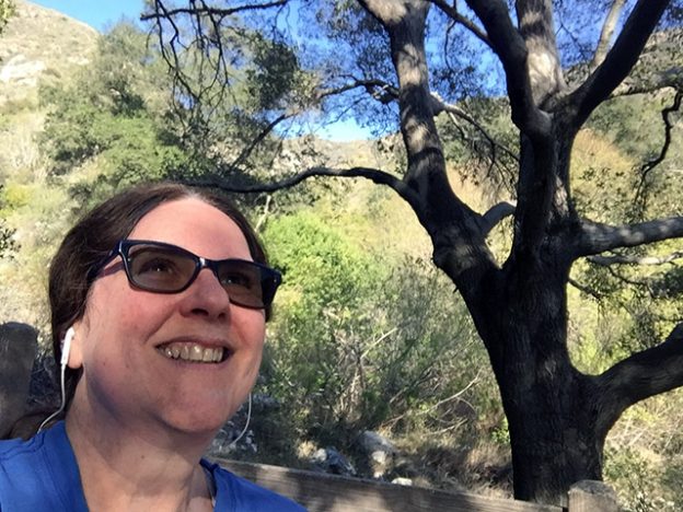 Laurie wearing light sun glasses and smiling mightily while sitting on a wooden bench under an oak tree.