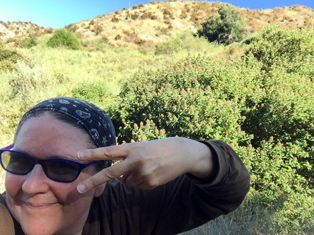 Laurie in a dark headscarf and shades dancing with her fingers making the peace sign while in the green meadow at the top of the hiking trail under blue skies.