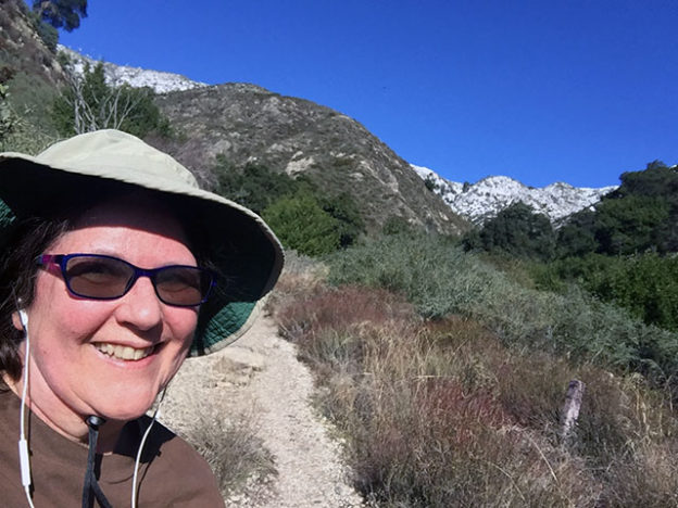 Laurie in hiking hat and sun glasses wearing a long sleeved brown sweatshirt smiles on the trail with the snow capped peaks visible behind her.