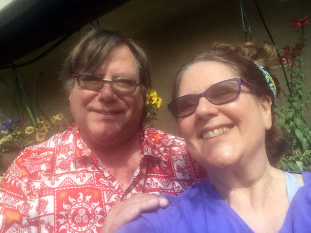 Mark in a red Hawaiian shirt with his arm around Laurie who is wearing a purple t-shirt in their garden by the front door of their home.