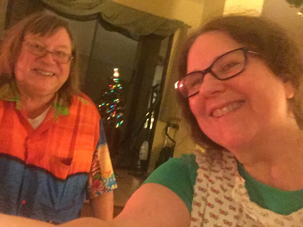 Laurie in an apron sits by Mark in his Hawaiian shirt and REALLY long hair while the Christmas Tree is reflected at night in the window behind them.